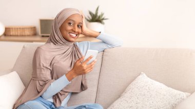 Başörtülü genç siyahi kadın iş için akıllı telefon kullanıyor ya da evdeki kanepede ders çalışıyor. Mutlu Müslüman kadın internette geziniyor, sohbet ediyor veya sosyal medyada cep telefonuyla paylaşıyor.