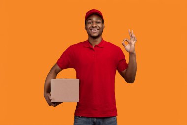 En İyi Teslimat Hizmeti Konsepti. Kırmızı şapkalı ve üniformalı gülümseyen siyah kurye elinde karton kutu ve sarı turuncu stüdyo arka planında izole bir şekilde duran işaret işareti gösteriyor.