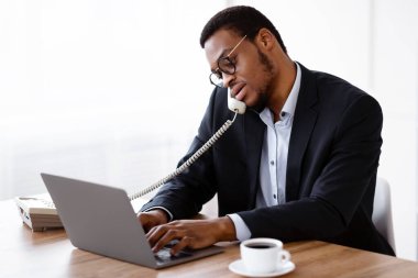 Siyahi kriz yöneticisi müşteriyle ya da meslektaşıyla telefonda konuşuyor ve modern dizüstü bilgisayarını kullanıyor, internetten verileri kontrol ediyor çalışma masasında kahve içiyor, ofis içi boş.