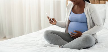 Gülümseyen siyah hamile kadın evde yatağında akıllı telefonuyla oturuyor, heyecanla cep telefonu ekranına bakıyor, mutlu Afrikalı kadın gebelik uygulaması ya da cep telefonu teklifi alıyor,