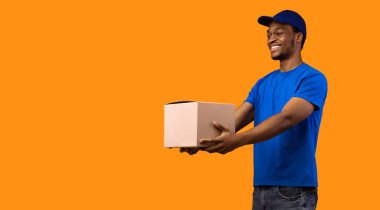 Teslimat servisi konsepti. Mavi şapkalı ve üniformalı gülümseyen siyah erkek kurye karton kutuyu tutuyor ve yan tarafa veriyor, kopyalama alanı, afiş. Sarı stüdyo arkasında duran adam.