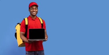 Afro-Amerikan kurye elinde boş ekranla dizüstü bilgisayar tutuyor, kırmızı üniforma ve termal çanta giyiyor, mavi stüdyo arka planında, panorama, afiş, kopyalama alanı üzerinde tek başına duruyor.