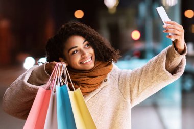 Alışveriş zamanı geldi. Mutlu Afro-Amerikalı kadın selfie çekiyor merkezde renkli çantalarla duruyor, dışarıda yürüyor, cep telefonunun kamerasına poz veriyor. Eğlence Konsepti