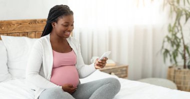 Mutlu Afrikalı genç hamile kadın evde akıllı telefonuyla rahatlıyor, sosyal ağlara bakıyor ya da hamilelik uygulamasını kullanıyor, yatak odasında oturuyor ve karnına dokunuyor,