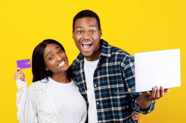 İnternetten alışveriş, kara cuma ve parayla ödeme. Mutlu, gülen, ağzı açık, genç Afrikalı Amerikalı bir adam dizüstü bilgisayar tutuyor, kadın kredi kartı gösteriyor, sarı arka planda izole edilmiş, stüdyo fotoğrafı.