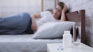 Bir kavanoz hap ve komodinin üzerinde bir bardak su ve arka planda yatan depresyonlu genç bir kadın, kopyalama alanı. Seçici odaklanma. İntihar ve uykusuzluk sorunu kavramı