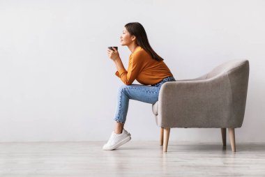 Genç Asyalı bir kadının koltukta oturup sıcak aromatik kahve içmesi, beyaz stüdyo duvarına yaslanıp dinlenmesi, boş yer. Güzel milenyum hanımı Tembel Gününde sıcak içeceğin tadını çıkarıyor