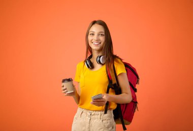 Sırt çantalı, cep telefonlu, elinde kahve olan mutlu genç bir kadın. Kameraya gülümsüyor, sarı arka plan. Mobil uygulama şehir rehberi kullanan ve çay içen bir kadın gezgin.