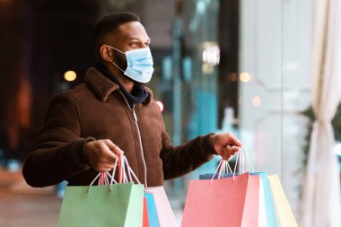 Tıbbi yüz maskesi takmış Afro-Amerikan bir adamın portresi alışveriş merkezinin vitrinine bakıyor, akşamları alışveriş merkezinin yanında elinde poşetlerle yürüyor. Kış Hediyeleri, Satış ve İndirim, Perakende