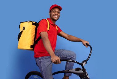 Hızlı Teslimat Servisi. Kırmızı şapkalı ve üniformalı gülümseyen Afro-Amerikan kurye bisiklet sürüyor ve mavi stüdyo arka planında izole edilmiş sarı termo çanta giyiyor. Sevkiyat ve posta