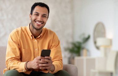 Mutlu Gülümseyen Adam telefonuyla internette geziniyor. Evdeki koltukta oturuyor. Akıllı Telefon için Neşeli Cep Telefonu Kullanıcı Deneme Yeni Uygulaması, Kameraya Bakma, Beleş Kopyalama Boşluğu, Model Uygulaması