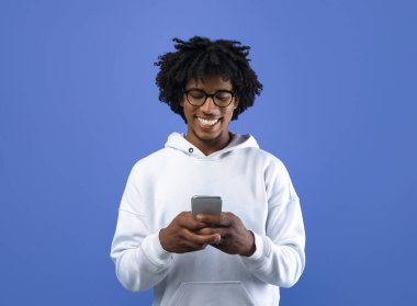 Mavi stüdyo arka planında yeni uygulamalar kullanarak cep telefonu ekranına bakan mutlu siyah bir genç. Akıllı telefon kullanan neşeli Afrikalı Amerikalı ergen, sosyal ağları tarıyor