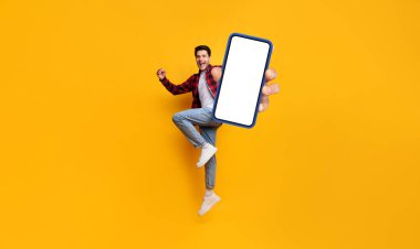Öyle mi? Neşeli adamın tüm vücut boyu portresi zıplıyor, yumruğunu sıkarak kazanan bir jest yapıyor ve cep telefonunu beyaz ekranda mobil uygulama için ücretsiz olarak gösteriyor, sarı arka plan
