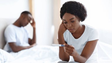 Depresyonda olan siyah kadın, elinde pozitif hamilelik testi, üzgün Afrikalı Amerikalı erkek, arka planda kafasına dokunuyor, istenmeyen hamilelik kavramı, kopyalama alanı.