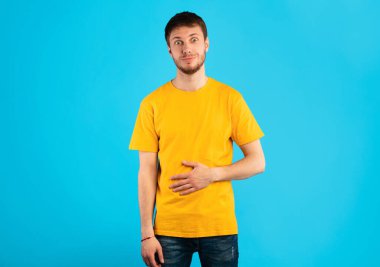 Midende Hoş Bir His. Tatmin olmuş gülümseyen genç adam ellerini karnına koyuyor, lezzetli bir yemek yedikten sonra keyfi yerinde oluyor, mavi arka planda tamamen soyutlandığını gösteriyor.