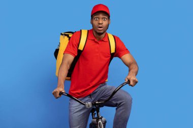 Hızlı Teslimat Servisi. Kırmızı şapkalı ve üniformalı komik Afro-Amerikan erkek kurye aceleyle bisiklete biniyor ve mavi arka planda ısıl sırt çantası takıyor.