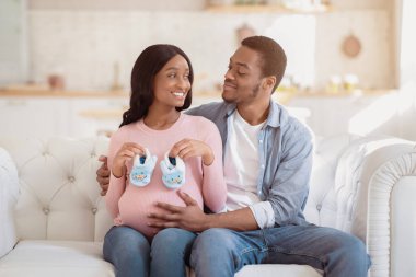 Mutlu siyah çift bebek bekliyor, kanepede birlikte oturuyorlar ve ellerinde küçük çocuk ayakkabıları var. Afrikalı Amerikalı sevgilisini seven, hamile sevgilisinin karnına dokunan.