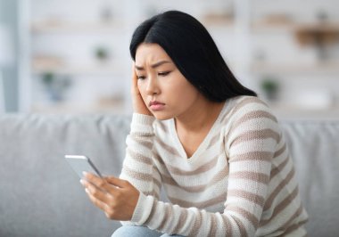Endişeli Asyalı kadın evde koltukta otururken Smartphone ekranına bakıyor, Hüsrana uğramış Koreli kadın cep telefonu tutuyor, kötü haber alıyor ya da anlamsız mesajlar okuyor, yakın çekim, boş alan