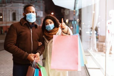 Hadi bunu alalım. Afro-Amerikan kadın portresi, tıbbi maske takıyor, vitrinlere bakıyor, akşamları alışveriş merkezinin yakınında kocasıyla birlikte çantalarla yürüyor. Mevsimlik Satış ve İndirim