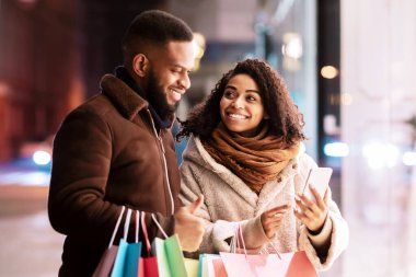 Mevcut Seçiliyor. Gülümseyen Afro-Amerikan kadın cep telefonunu erkeğe gösteriyor, cihazı işaret ediyor, alışveriş torbaları tutan adam, akşamları dışarıda duruyor. Teklif, Perakende ve Satın Al
