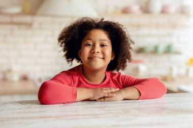 Mutfakta masada oturan kıvırcık siyah kızın yakın plan portresi, evde dinlenen Afro-Amerikan kadın çocuk, kreş çocuğu kameraya bakıyor ve gülümsüyor.
