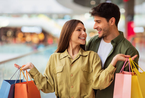 Вместе ходим по магазинам. Счастливая жена держит в руках разноцветные сумки покупателя, чтобы муж стоял в современном гипермаркете. Клиенты в торговом центре наслаждаются покупкой одежды по выходным. Концепция сезонных продаж