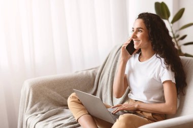 Uzun kıvırcık saçlı, mutlu esmer kadın hafta sonunu evde geçiriyor, dizüstü bilgisayarla koltukta oturuyor ve cep telefonuyla sohbet ediyor, neşeli bir şekilde boş bir yere bakıyor.