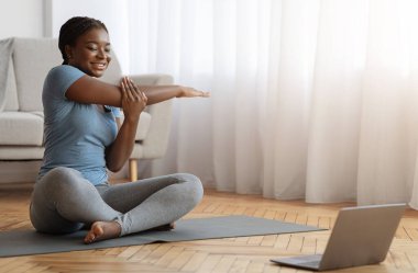 Neşeli Afrikalı kadın dizüstü bilgisayarın önünde geriniyor, ev ödevi yapıyor, ev içi spor için ısınıyor oturma odasında spor yapıyor, oturma odasında spor yapıyor, boş alan arıyor.