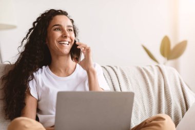 Neşeli, serbest çalışan genç bir kadın cep telefonuyla konuşuyor ve dizüstü bilgisayar kullanıyor, izolasyon sırasında evden çalışıyor, kopyalama alanına bakıyor. Mutlu kıvırcık kadın telefonda konuşuyor.