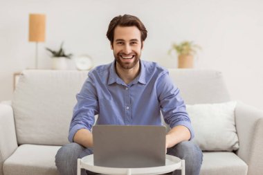 Dizüstü bilgisayardaki mutlu adam internette çalışıyor ve evdeki koltukta oturan kameraya gülümsüyor. Uzaktan kumandalı başarılı adam içeride çalışıyor. Serbest meslek hayatı konsepti