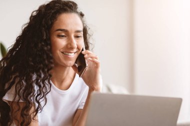 Cep telefonuyla konuşan, dizüstü bilgisayar kullanan, izolasyon sırasında evden çalışan, serbest çalışan mutlu genç bir kadın. Kıvırcık kadın telefonda konuşuyor ve bilgisayar ekranına bakıyor.