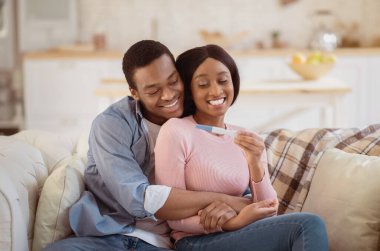 Güzel siyah çift pozitif hamilelik testi yapıyorlar ve evdeki kanepede kucaklaşıyorlar. Genç Afrikalı Amerikalı adam mutlu karısını kucaklıyor, bebeğini öğrendiği için mutlu.