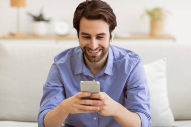 Neşeli adam akıllı telefon ve kulaklık kullanarak video görüşmesi yapıyor. Evdeki koltukta oturuyor. Oturma odasında gezici oyun oynayan adam. Gadget, Yeni Uygulama Kavramı