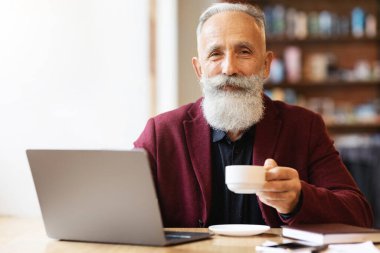 Yakışıklı yaşlı adam kafede vakit geçiriyor, kahve ya da çay içiyor ve dizüstü bilgisayar kullanıyor, web sörfü yapıyor ya da internette çalışıyor, kameraya gülümsüyor, boş bir alanda. Emeklilik konsepti sırasında aktif yaşam tarzı