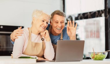 Modern mutfakta oturan dizüstü bilgisayarlı mutlu son sınıf çifti videoları. Yaşlı eşler içeride ekranlara el sallarken görüntülü arama yapıyorlar. Çevrimiçi İletişim