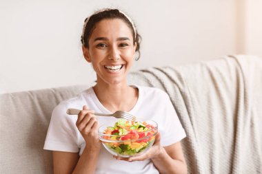 Mutlu bayanın elinde taze salata ve çatal, kameraya gülümseyen, evdeki kanepede oturan, fotokopi çeken. Sağlıklı genç esmer kadın öğle yemeğinde sebze yiyor.