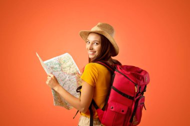 Sırt çantalı, elinde şehir haritası olan gülümseyen genç bir turist. Neşeli bayan haritasını tutuyor, omzunun üstünden bakıyor, büyük seyahat fırsatlarından etkileniyor, boşluğu kopyalıyor.