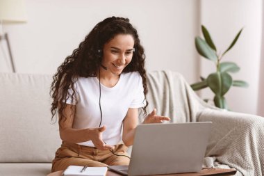 Kulaklıklı neşeli genç bir kadın online konferans yapıyor, evden çalışıyor, fotokopi çekiyor. Arkadaşlarıyla ya da sevgilileriyle video görüşmesi yapan mutlu kıvırcık kadın, dizüstü bilgisayarını, oturma odasını kullanıyor.