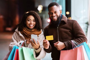 Ticaret ve Kolay Ödeme Kavramı. Mutlu Afro-Amerikalı erkek ve kadın akıllı telefon kullanıyor, bankamatik kartı gösteriyor ve alışveriş çantalarını tutuyor, akşamları dışarıda duruyor, kameralara bakıyor.