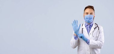 Kendine güvenen maskeli genç erkek doktor gri stüdyo geçmişine eldiven takıyor. Koronavirüs salgını sırasında önlük giyen başarılı bir tıp çalışanı.