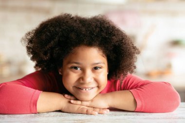 Evdeki mutfakta poz verirken gülümseyen tatlı siyah kıvırcık kız portresi, masaya yaslanan sevimli Afro-Amerikan bayan çocuk, ellerinin üzerinde dinlenen kafa,