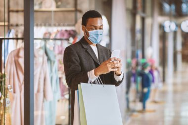 Düşük fiyatlar, indirimler ve mobil uygulama satışları ve koronavirüs salgını sırasında modern cihazlar. Milenyum Afrika kökenli koruyucu maskeli Amerikalı birçok renkli paketi olan adam vitrinin yanındaki telefona bakıyor.