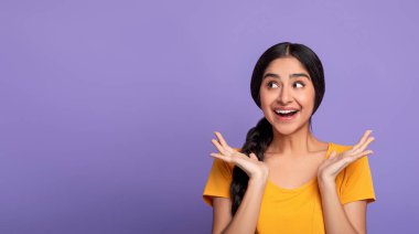 Omg. Coşkulu genç Hintli kadının portresi ellerini açıyor ve reklam ya da promosyon metinleri için bir kenara bakıyor, mor arka plan, stüdyo çekimi, afiş
