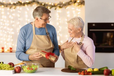Elderly Husband Giving Wrapped Gift To Wife Celebrating Christmas Holiday In Kitchen At Home. Happy Family Xmas Celebration, Winter Holidays Season Concept. Senior Couple Preparing X-Mas Dinner