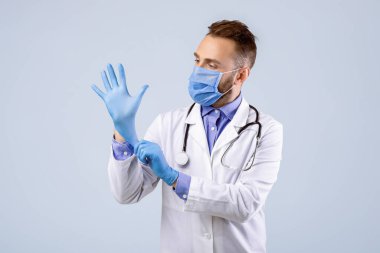 Tıbbi maskeli profesyonel bir doktor gri stüdyo geçmişine eldiven takıyor. Covid-19 salgını sırasında önlük giyen kendine güvenen bir doktor. Viral hastalıkların önlenmesi