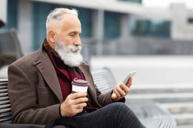 Gri saçlı, sıcak giyimli, gezici foe kullanan ve sokakta otururken kahve içen, boş zamanlarının tadını çıkaran, sosyal medyayı kontrol eden ya da sohbet eden, boşluğu kopyalayan yaşlı bir adam.