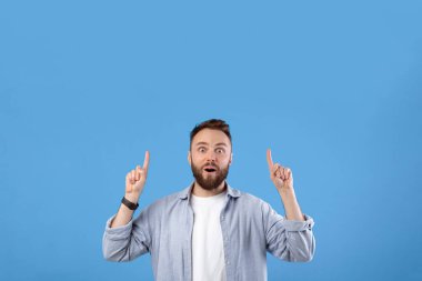 Yakışıklı sakallı, şok olmuş yüz ifadesi olan, mavi stüdyo arka planında boş bir yeri işaret eden adam. Milenyum adamı ürününüzü tanıtmak için büyük indirimler sunuyor.