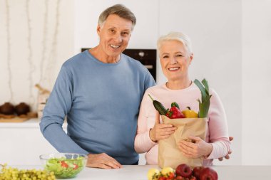 Evde, modern mutfakta birlikte duran sebzeli sebze dolu alışveriş torbası taşıyan mutlu yaşlı çift. Market alışverişinden sonra evde poz veren yaşlı eşler, süpermarket yiyecek teslimatı