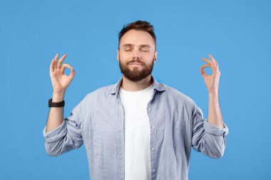 Stres yönetimi konsepti. Huzurlu genç adam gözleri kapalı meditasyon yapıyor, elleri mavi stüdyo geçmişiyle Gyan Mudra yapıyor. Milenyum adamı sakinliğini koruyor, iç dengeyi buluyor.