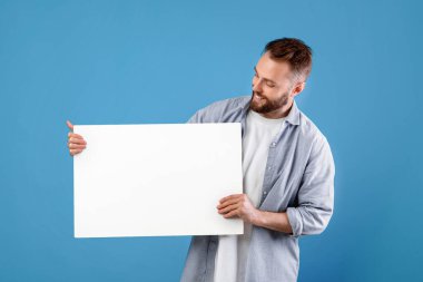 Mavi stüdyo arka planında beyaz boş plaket sergiliyor. Logo ya da tasarım için maket yapıyor. Boş reklam pankartı olan olumlu milenyum adamı, boşluğu kopyala